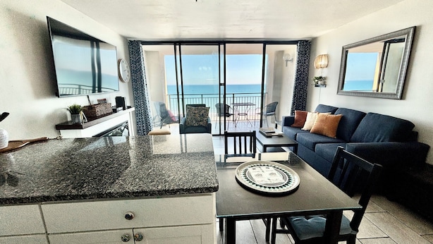 A modern living room with a kitchen area, a dining table, and a sofa. There is a large window providing a view of the ocean. - Perfect Peace:Spacious 1 BR King, Walk-in Shower, Big Balcony, Undeveloped Beach (Myrtle Beach)