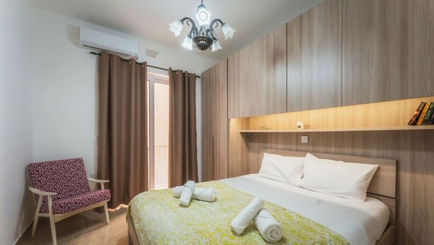 A hotel room with a bed, a chair, a ceiling light, and a wooden panel wall. - Marsalforn Apartment Walk to Beach Seaside Living (Żebbuġ)
