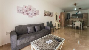 A living room with a grey sofa, a coffee table with a patterned top, and a dining area with a table and chairs. - Marsalforn Apartment Walk to Beach Seaside Living (Żebbuġ)