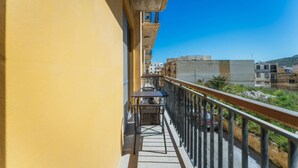 A balcony with a table and chairs, a metal railing, and a view of buildings and greenery. - Marsalforn Apartment Walk to Beach Seaside Living (Żebbuġ)