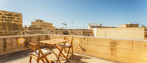 A rooftop terrace with a table and chairs, a view of surrounding buildings, and a clear sky.