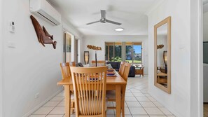 A dining area with a wooden table and chairs, a ceiling fan, and a view of the outdoors through large windows. - Oceania by Experience Jervis Bay (Vincentia)