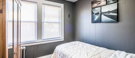 A bedroom with a bed, a radiator, a window with blinds, and wall-mounted artwork.