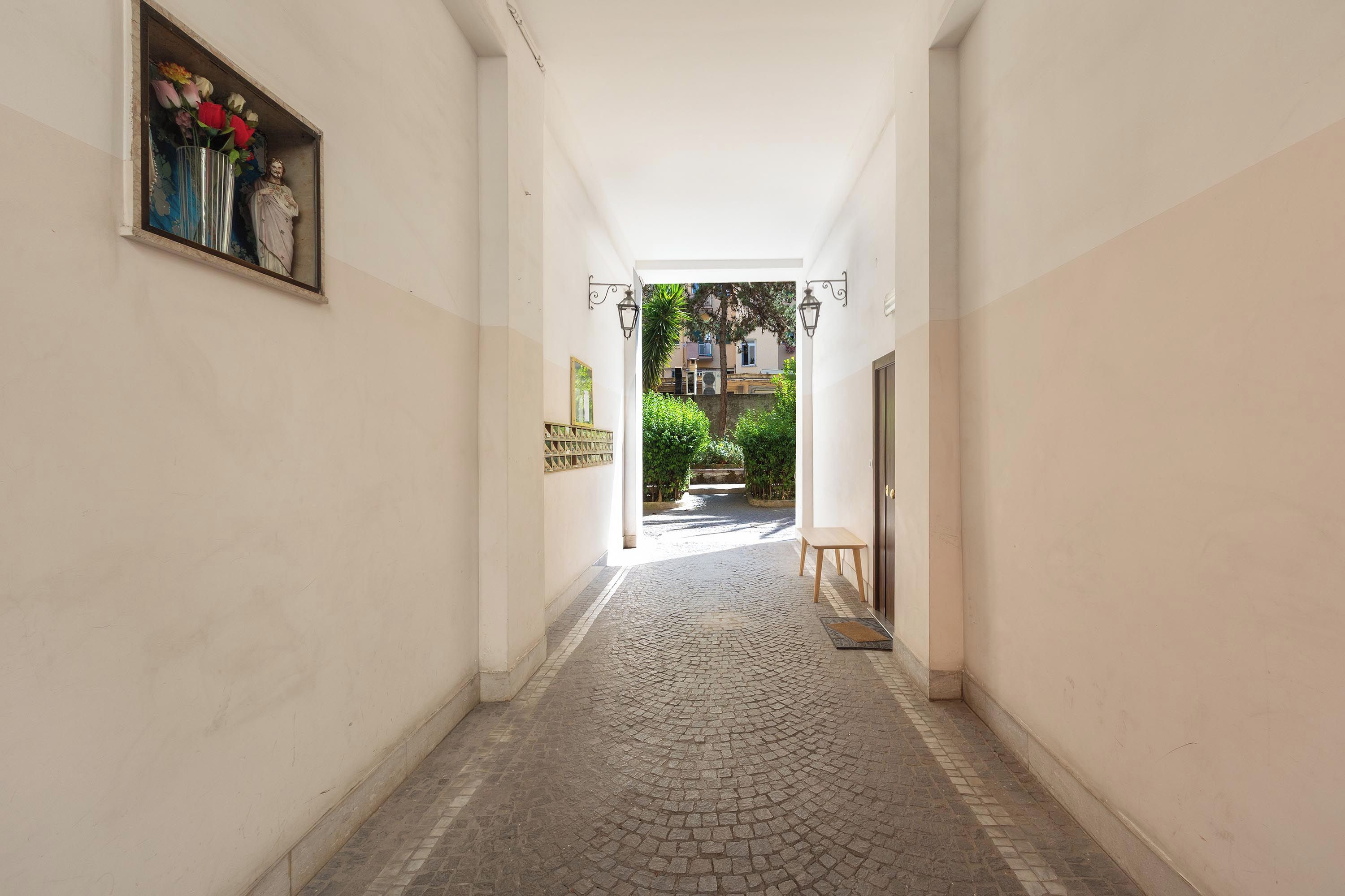 A corridor with a tiled floor leading to an outdoor area with greenery and buildings in the background.