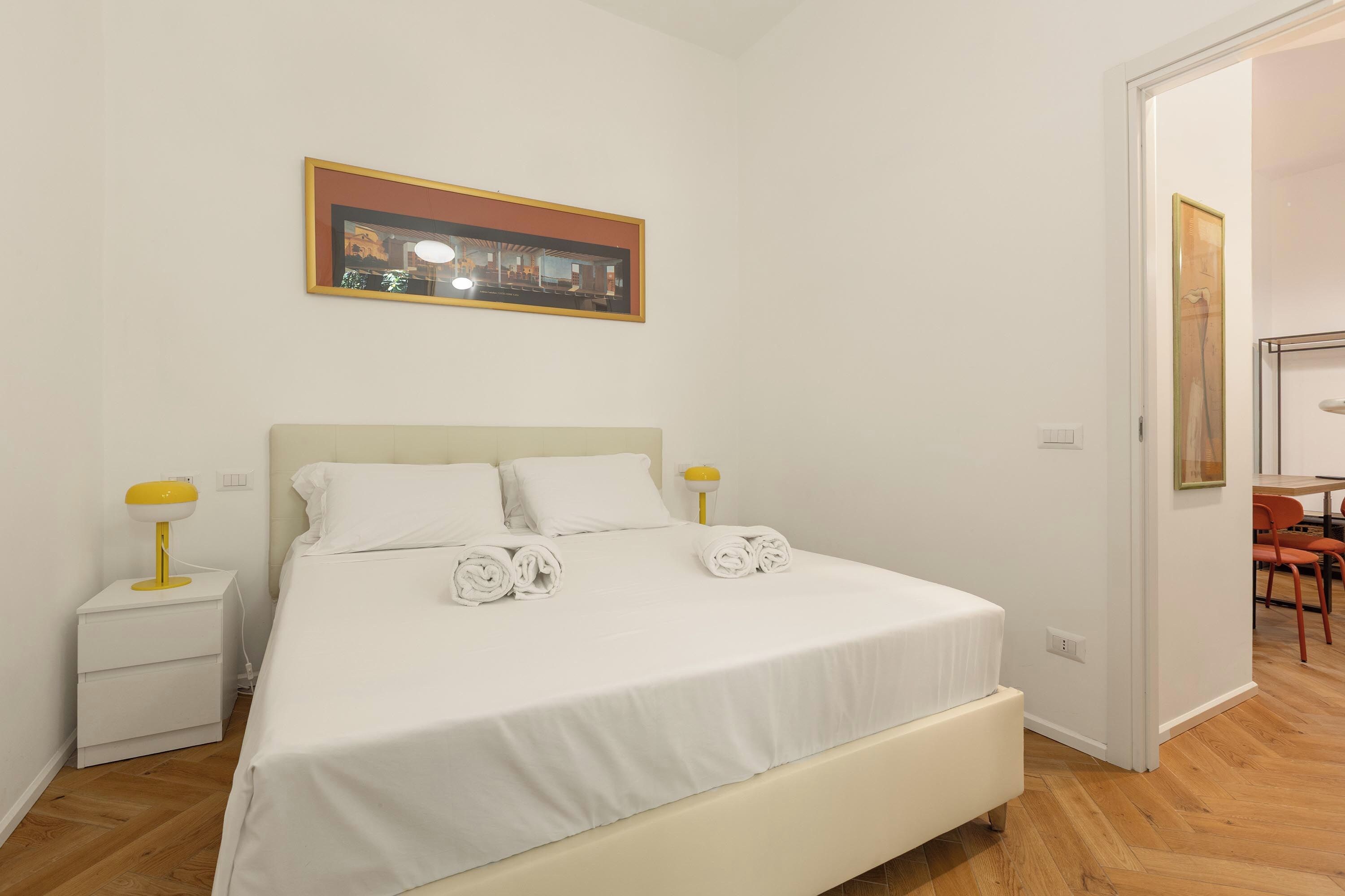 Ein Hotelzimmer mit einem Einzelbett, einem Nachttisch, einer Lampe und einem gerahmten Bild an der Wand.