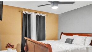 Un dormitorio con una cama de madera, ropa de cama blanca, un ventilador de techo y una ventana con cortinas blanco y negro.