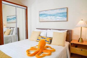 Ein Hotelzimmer mit einem Bett, einem Nachttisch, einer Lampe, einem Bild vom Meer und einer großen Plüsch-Tintenfischfigur auf dem Bett.