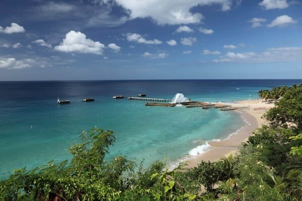 Blick auf die Küste mit einem Steg, der sich in türkisfarbenes Wasser erstreckt, einem Sandstrand und üppiger Vegetation.