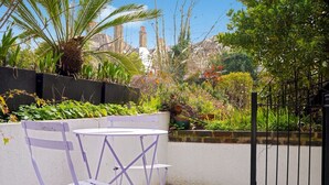 Un patio con un tavolo bianco e sedie viola, circondato da vegetazione lussureggiante e un muro in mattoni.
