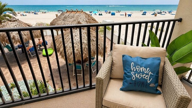 A balcony with a wicker chair, a blue pillow with 'home sweet home' text, and a view of a beach with umbrellas and people. - Beachfront Getaway with Resort-Style Pool (Madeira Beach)