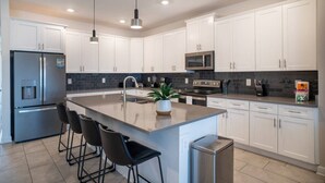 A modern kitchen with white cabinets, stainless steel appliances, a central island with bar stools, and pendant lighting. - 6BR Villa w Pool & Game Room near Disney (Davenport)