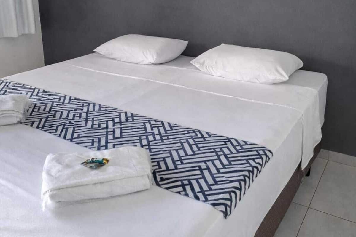 A neatly made bed with white linens and a geometric-patterned throw, a gray wall with two framed floral artworks, and a white curtain on the left.