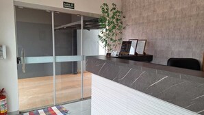 Una zona de recepción con puerta de vidrio marcada 'SAÍDA DE EMERGENCIA', mostrador de recepción, planta en maceta y extintor.