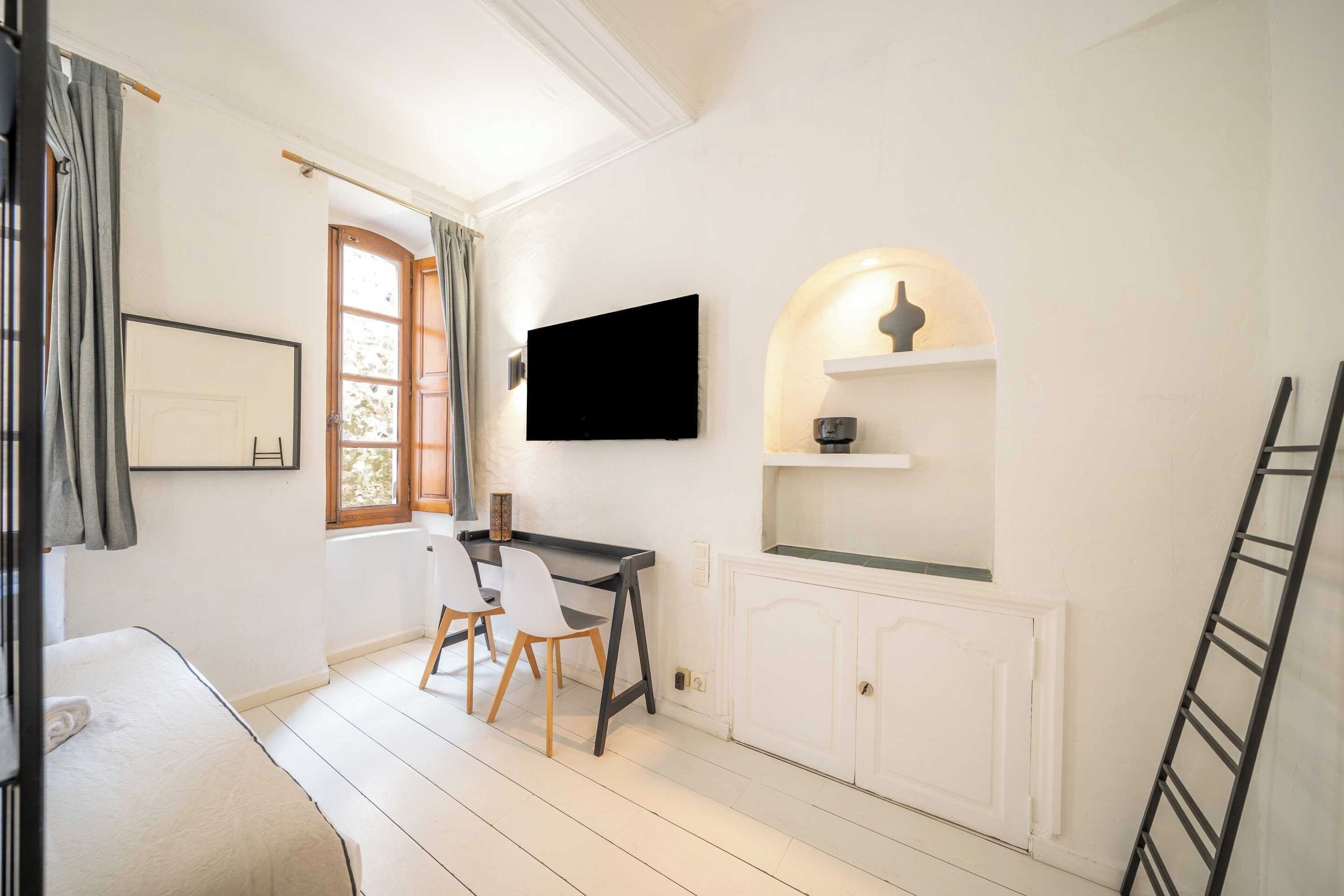 Een kamer met een bed, een bureau met stoelen, een televisie, een raam met gordijnen en een witte kast.