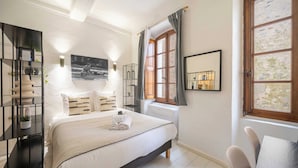 Un dormitorio con cama, mesita de noche, espejo, ventana con cortinas y estante fijado a la pared.