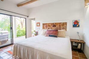 Een slaapkamer met een groot bed, een stenen hoofdbord, een glazen deur naar een terras en een klein nachtkastje met een lamp.