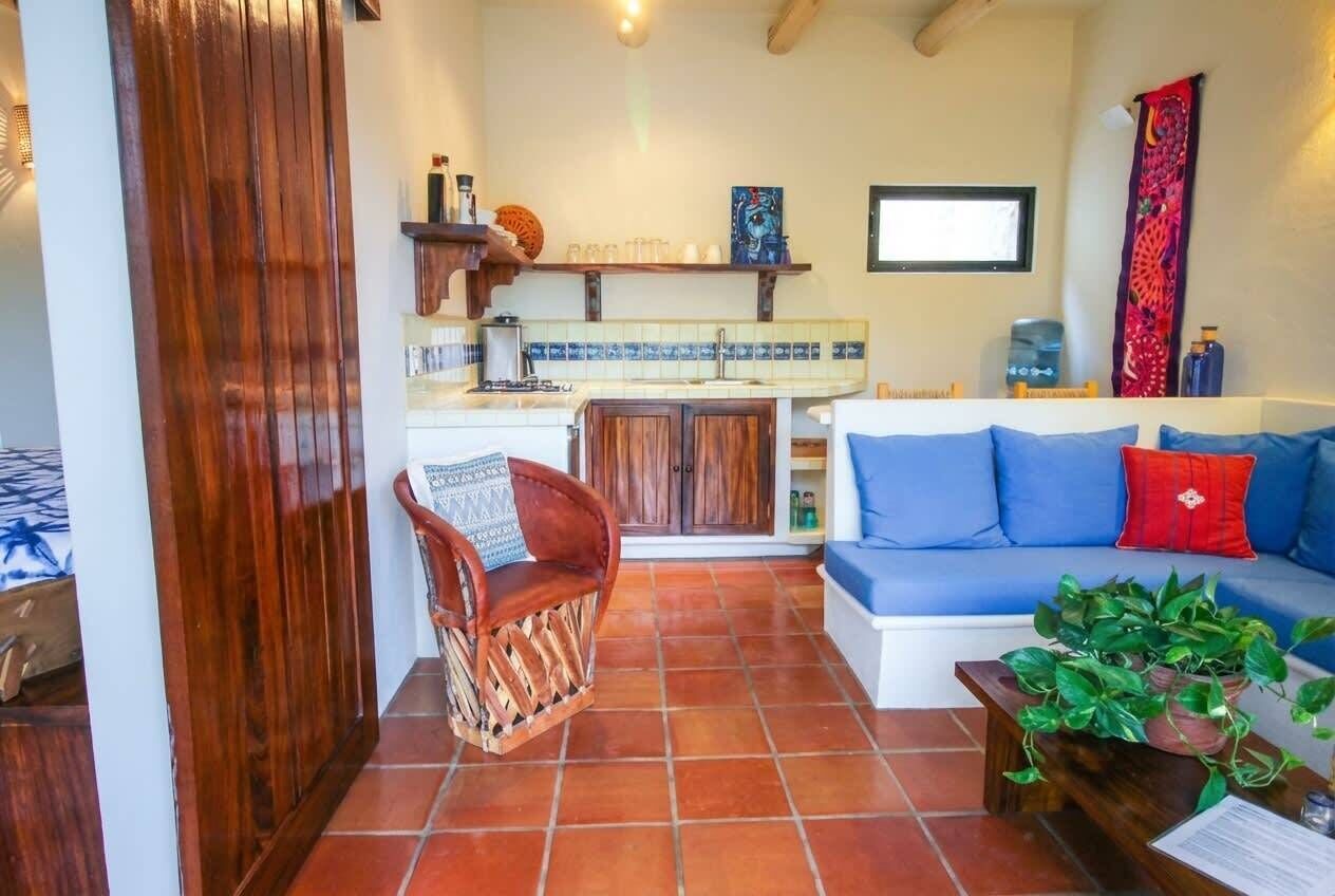 Una sala de estar acogedora con un sofá, una silla de madera, suelo de baldosas y una zona de cocina con fregadero y armarios.