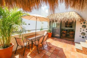 Een balkon met een tafel en stoelen, een potplant, een rieten parasol en uitzicht op het interieur.