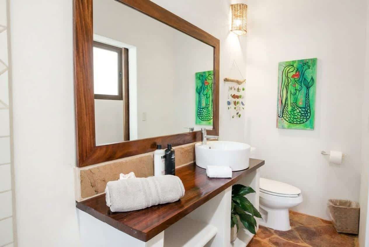 A bathroom with a wooden vanity, a white sink, a mirror, and a toilet. There are two green wall art pieces and a hanging light fixture.