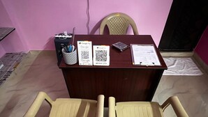 Una habitación con un escritorio de madera, dos sillas de plástico, una silla con cojín, una mesita con una taza y bolígrafos, y una pared con un código QR.
