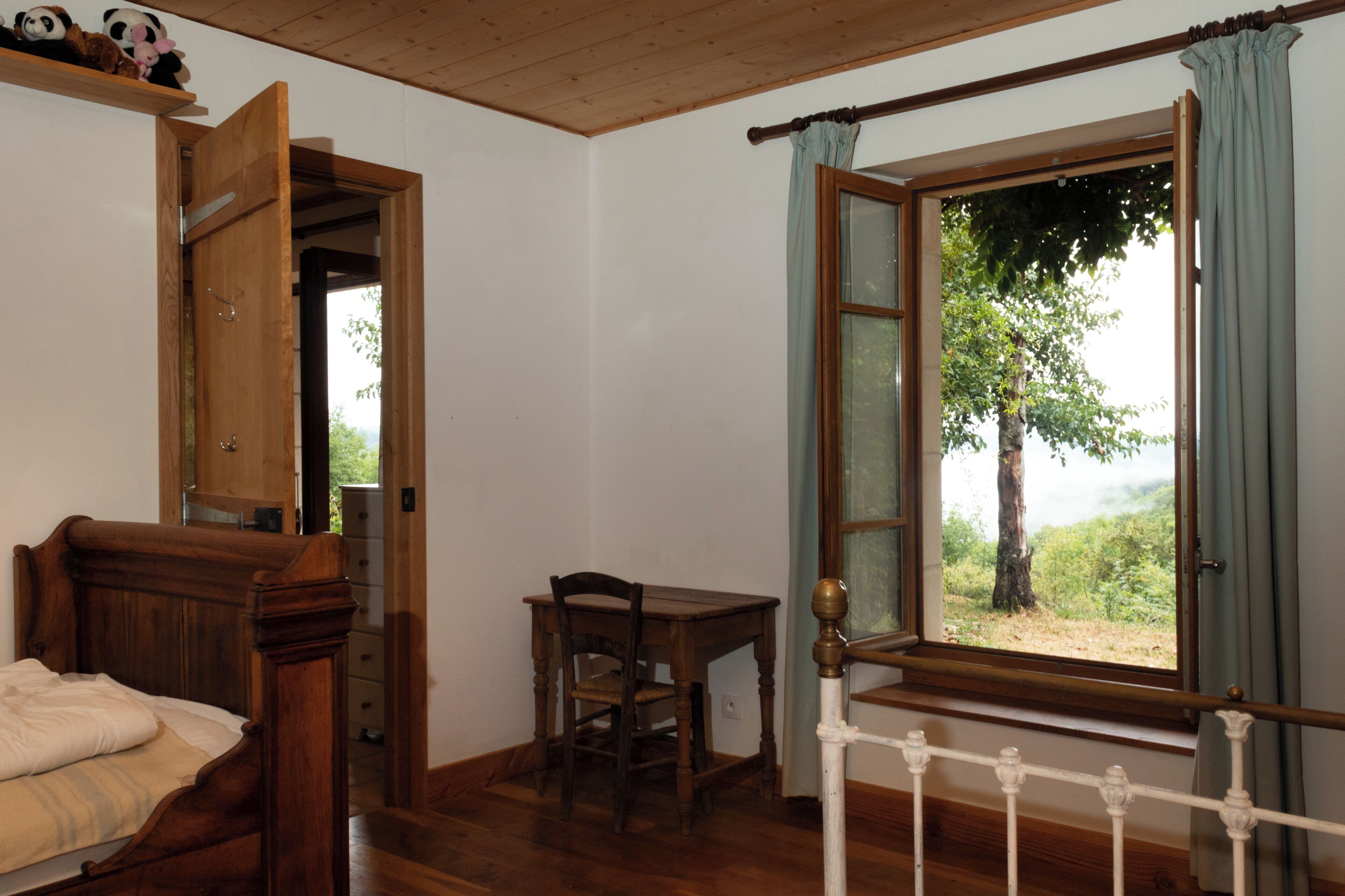 Ein Zimmer mit einem hölzernen Bett, einem Schreibtisch aus Holz, einem Stuhl, einem Fenster mit Vorhängen und einer Tür zu einem anderen Raum.