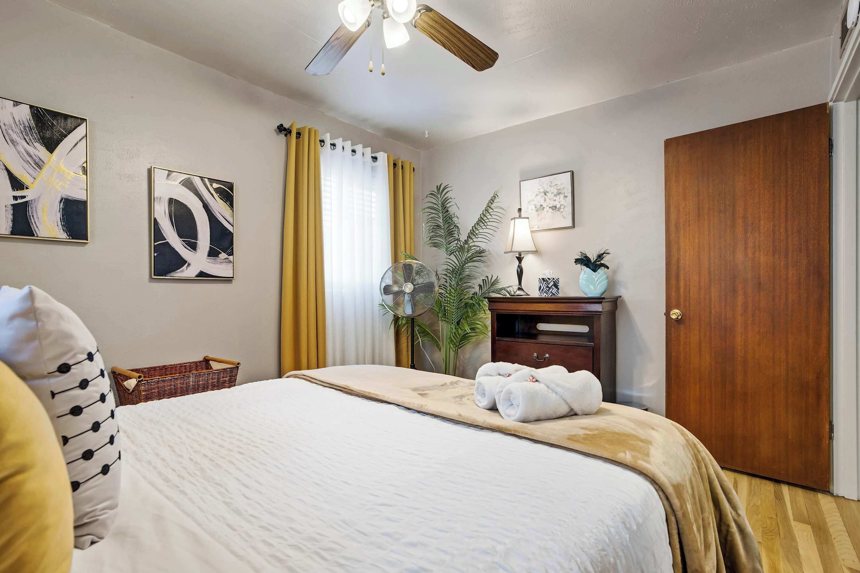 A hotel room with a bed, a ceiling fan, a wooden door, a plant, and artwork on the walls.