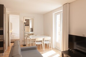 A modern living room with a sofa, a dining table with chairs, a TV, and a large window with curtains. - FLH Alegria Elegant Duplex (Lisbon)