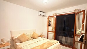 Un dormitorio con una cama grande, mesitas de noche, un equipo de aire acondicionado, una puerta corrediza de vidrio y una pequeña mesita de noche con lámpara.