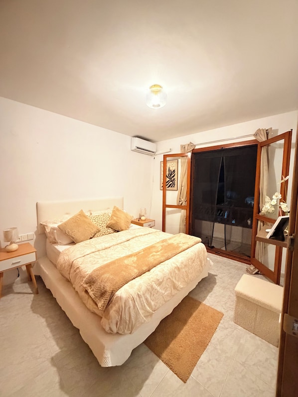 Une chambre dotée d’un grand lit, de tables de chevet, d’un appareil de climatisation, d’une porte-fenêtre coulissante et d’une petite table de chevet avec une lampe.