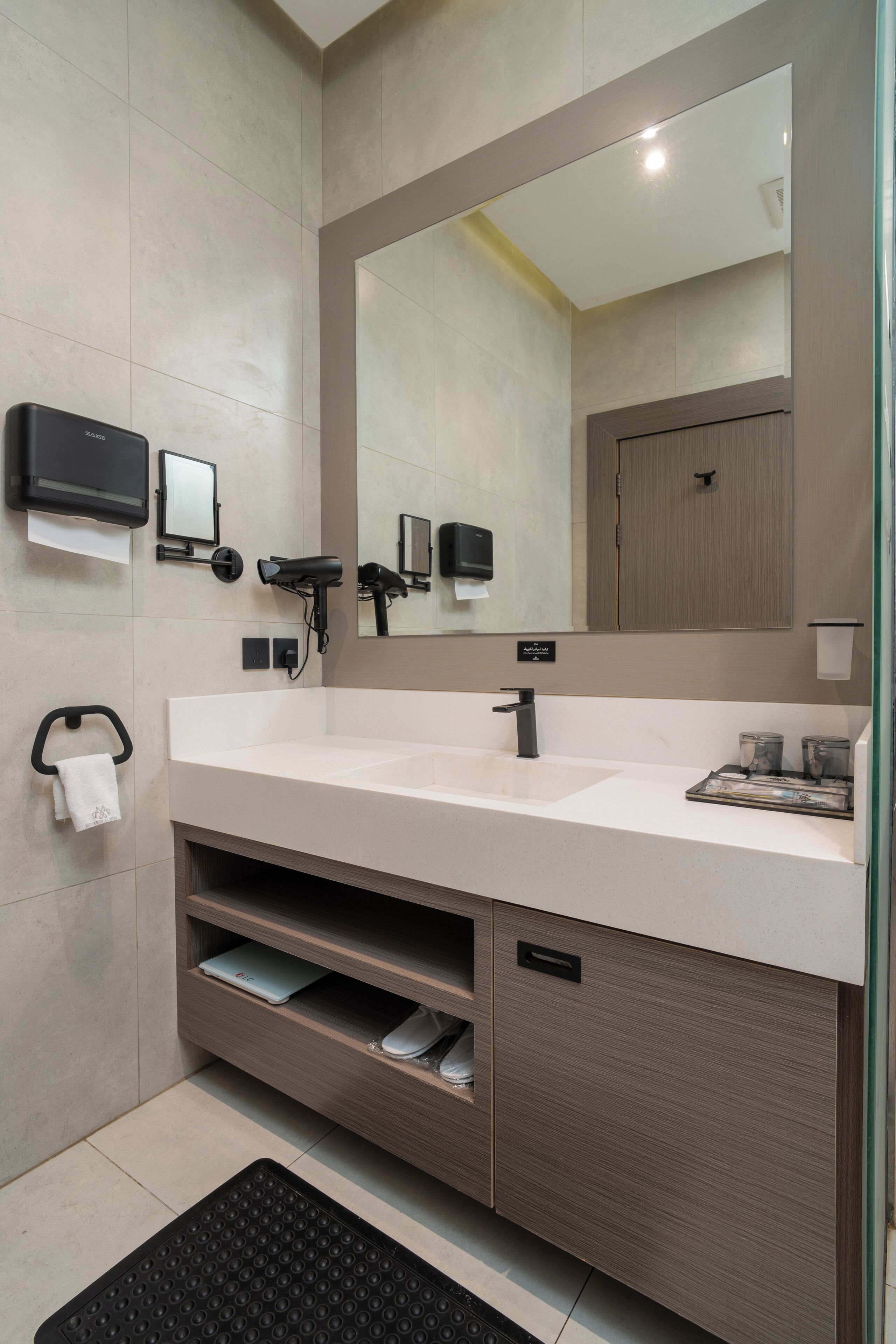 A modern bathroom with a large mirror, a white sink, a towel rack, a hairdryer, and a shelf with a laptop.