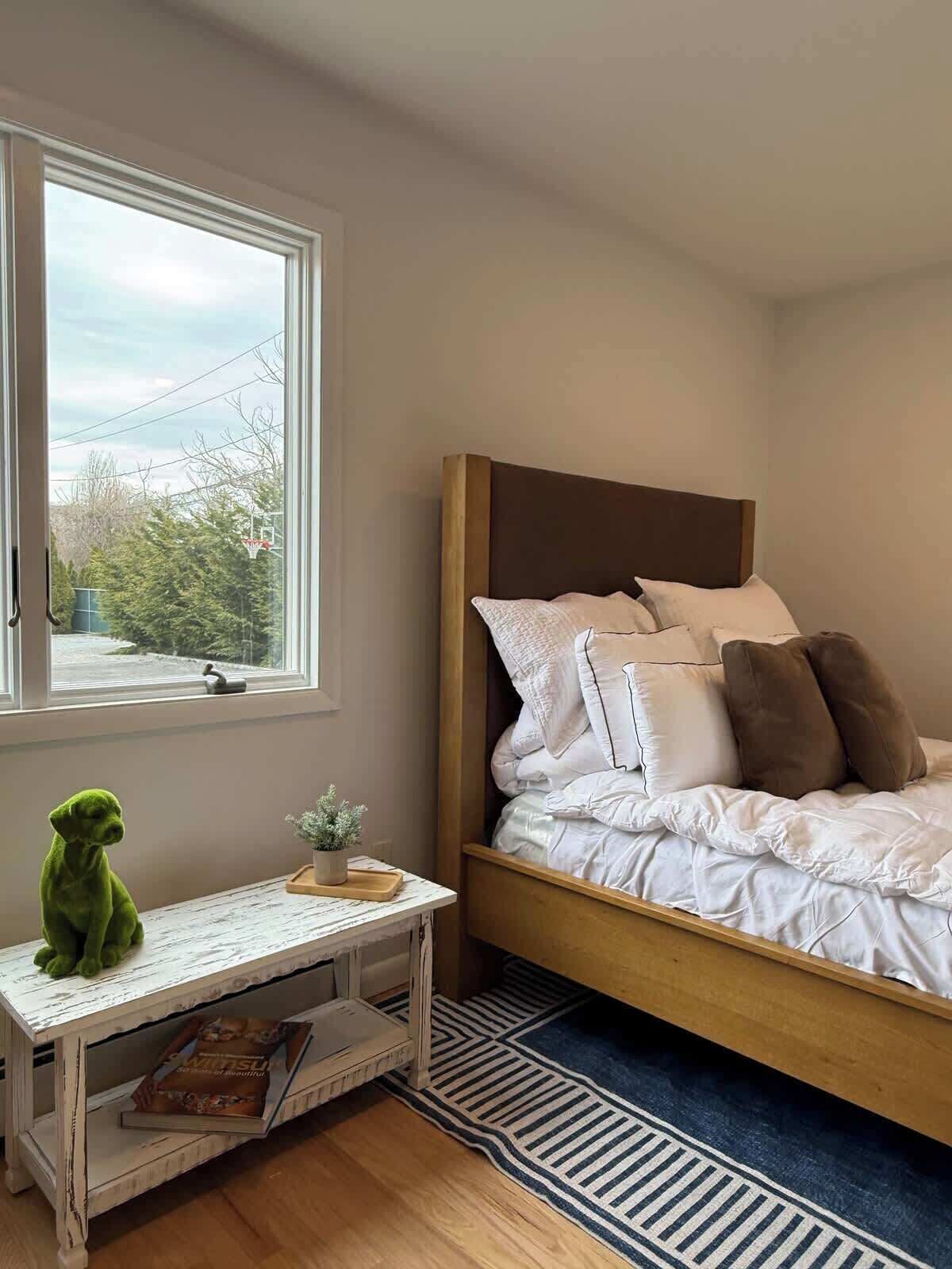 Ein Schlafzimmer mit einem Bett, einem kleinen Tisch mit einem grünen Plüschtier, einem Fenster und Blick auf Bäume sowie einen Basketballkorb.