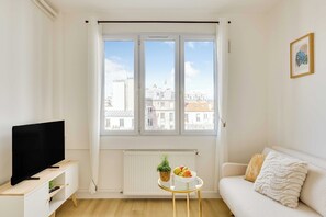 Un salon avec un téléviseur, un canapé, une petite table sur laquelle se trouvent des fruits, et une fenêtre donnant sur la ville.