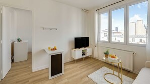 A modern, minimalist living room with a television, a small table with fruit, and a view of the city through large windows. - Cozy Apartment 1b/2p - Paris 14th (Paris)