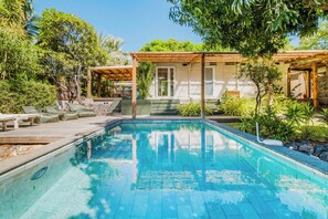 Ein Pool mit Holzterrasse, überdachtem Sitzbereich und üppiger, umgebender Bepflanzung.