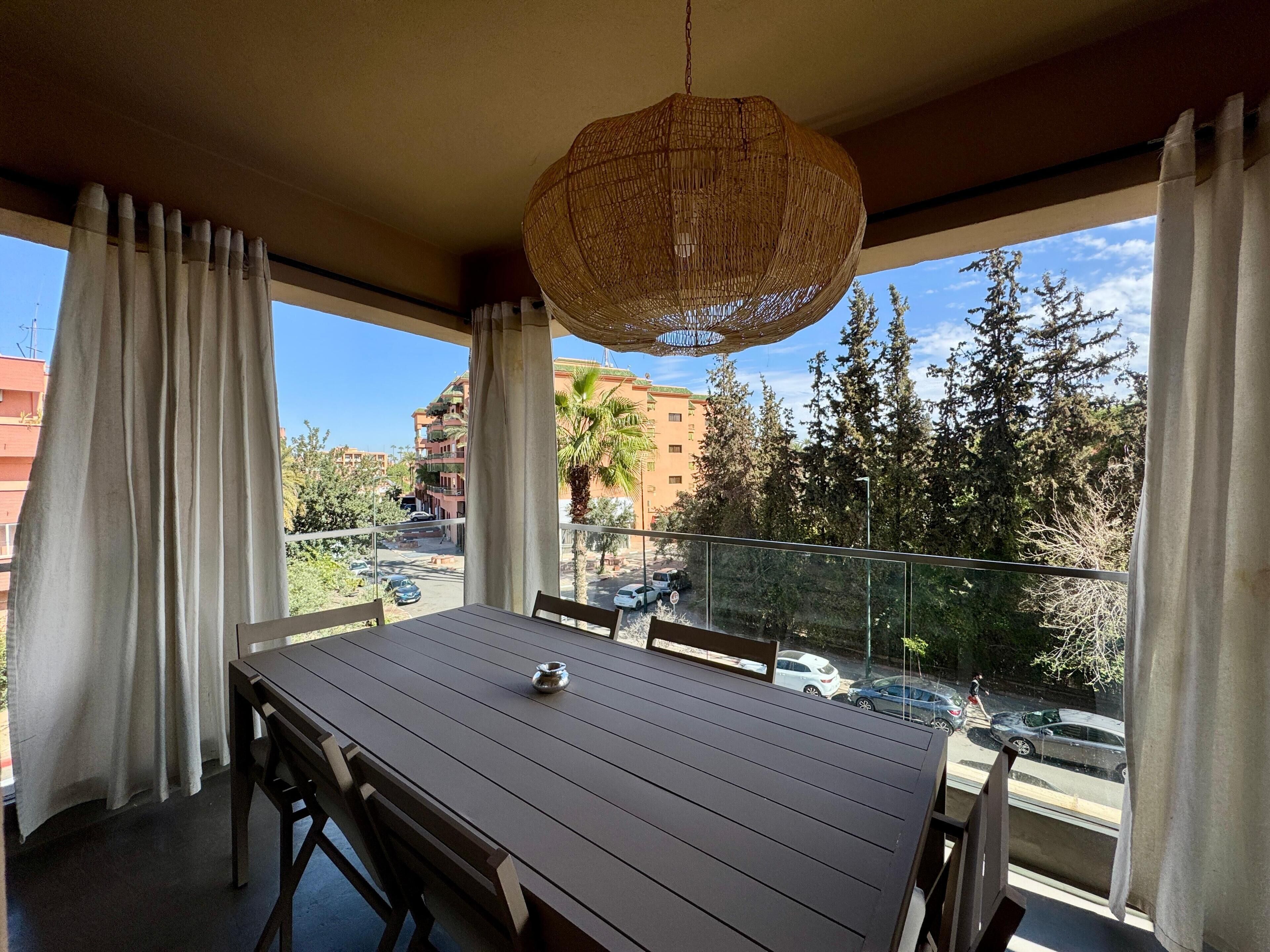 Un balcon avec une table et des chaises, un luminaire suspendu, et une vue sur la rue et les immeubles.