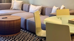 Een moderne woonkamer met een hoekbank, een ronde houten salontafel, een glazen eettafel en een wandsculptuur.