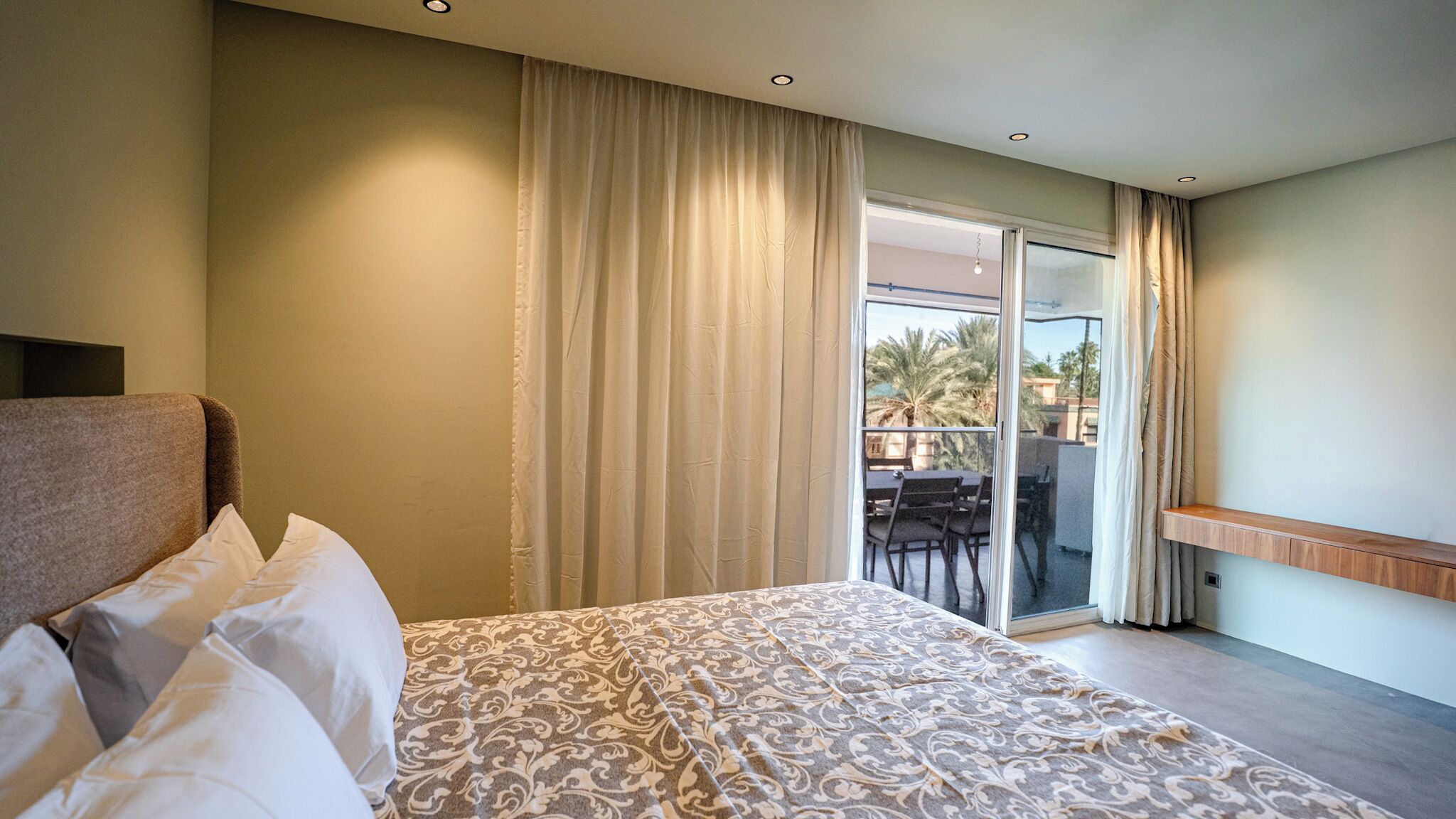 Une chambre d’hôtel avec un lit, un balcon doté d’une table et de chaises, et une vue sur des palmiers.