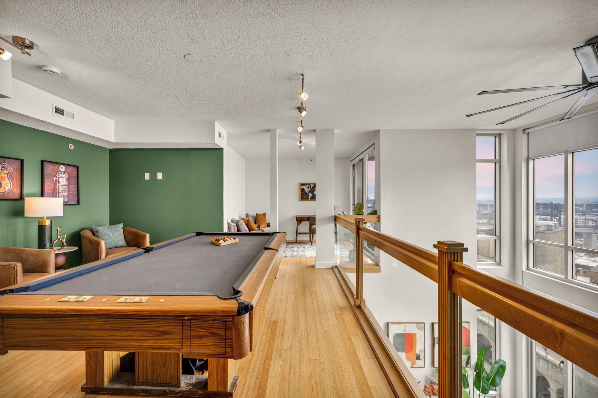 A pool table in a room with a view of the city through large windows, a staircase, and a green wall.