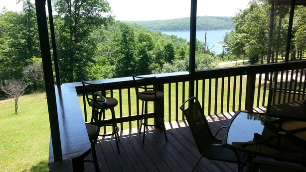 A wooden deck with a table and chairs, overlooking a lake and forest. - 4-bedroom house overlooking Lake Norfork with AC comfort (Mountain Home)