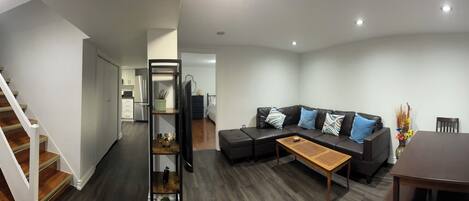 A modern living room with a sofa, coffee table, and a dining table. A staircase leads to a kitchen area.
