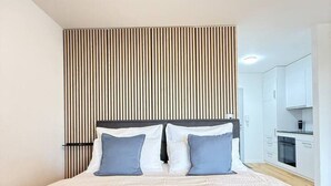 A modern bedroom with a large bed, a wooden slat wall, and a kitchenette in the background. - Lake view apartment with balcony (Rorschach)