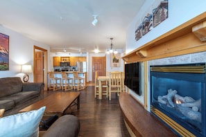 A living room with a fireplace, wooden furniture, and a kitchen in the background.