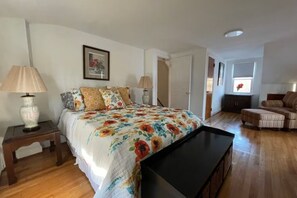 Een slaapkamer met een bed, bedlampjes, een houten nachtkastje, een zwarte tv-meubel en een bank, plus een raam met uitzicht.