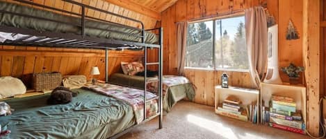 Une chambre dans une cabane en bois, avec des lits superposés, une petite fenêtre avec des rideaux, une table de chevet avec des livres et un sol recouvert d’une moquette.