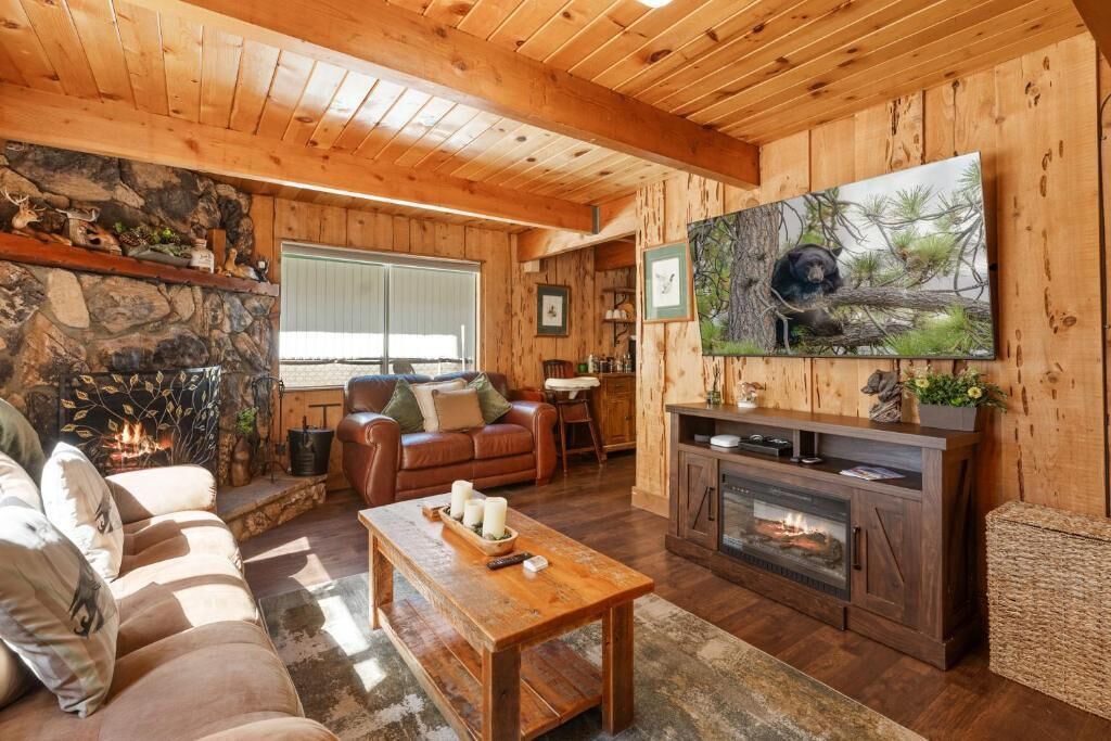 Un acogedor interior de cabaña con chimenea de piedra, techo de madera, televisor grande montado en la pared y un área de estar cómoda con sofá y mesa de centro.
