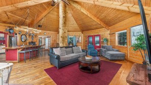 Un intérieur de chalet en bois spacieux, avec un plafond haut, un coin repas, un salon doté d’un canapé et de fauteuils, et un espace cuisine.