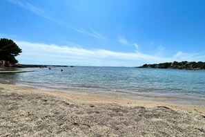 A beach with clear water, pebbly shore, and distant trees. - Le Petit Nid Azuré Studio AC Balcony Private Park (Saint-Raphaël)