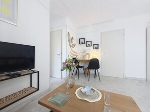 Ein modernes Wohnzimmer mit Flachbildfernseher, Couchtisch aus Holz, einer Vase mit Blumen und einem Essbereich mit Stühlen und Tisch.