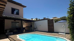 A swimming pool with a wooden deck, a covered patio area, and a view of a residential building. - Casa Arembepe - Camaçari-BA (Camaçari)