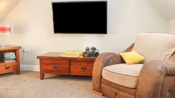 Una habitación acogedora con un televisor de pantalla plana montado en la pared, un sillón marrón con un cojín amarillo, una mesita de madera con libros y un juego de mesa, y una pequeña mesa de centro con una tetera y tazas.